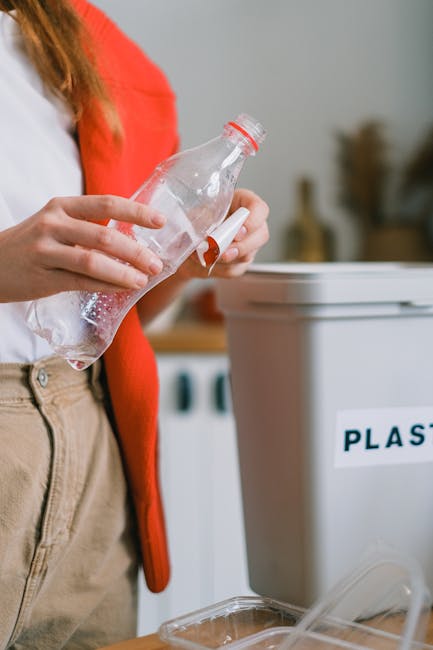 A person dressed in an orange jumpsuit and white shoes is standing indoors on a plain, smooth, grey floor. They are holding two large, blue, semi-transparent plastic rubbish bags filled with waste; th
