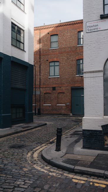 A narrow urban street scene featuring several multi-storey buildings with distinct architectural styles, including a prominent red-brick structure with arched windows and decorative stonework, alongsi